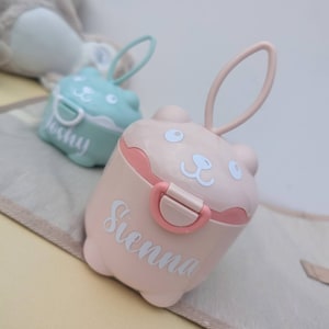 May include: Two pink and blue plastic containers with animal faces and lids. The containers have a handle and are labelled "Sienna" and "Joshy".