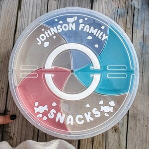 May include: A clear, circular snack container with a white handle, divided into five sections. The sections contain snacks of various colours. The lid is printed with the words "JOHNSON FAMILY" and "SNACKS" in white.