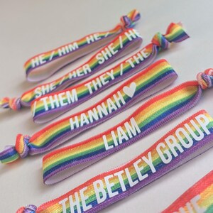 May include: Rainbow elastic hair ties with white text. The text on the hair ties reads "HE/HIM", "SHE/HER", "THEY/THE", "HANNAH", "LIAM", and "THE BETLEY GROUP".
