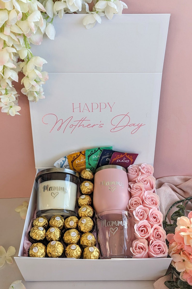 May include: A white gift box with the words "Happy Mother's Day" on the lid. The box contains a pink tumbler, a glass with a heart design, a candle, a box of chocolates, and a pack of tea bags. The box is surrounded by pink roses.