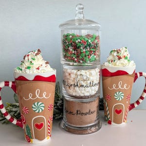 May include: A glass jar with three sections filled with cocoa powder, mini marshmallows, and red, green and white sprinkles. Two gingerbread-themed mugs with whipped cream and sprinkles are on either side of the jar.