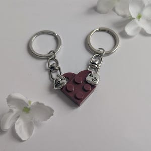 May include: Two matching keyrings featuring a heart-shaped charm made of maroon building blocks. Each keyring has a silver ring and a small heart charm with the letters "P" and "M". The keyrings are connected to form a complete heart.