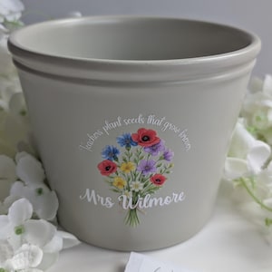 May include: A light grey ceramic plant pot featuring a colourful floral bouquet illustration in the centre. Above the flowers, the text reads "Teachers plant seeds that grow forever," and below the bouquet, the name "Mrs Wilmore" is printed.