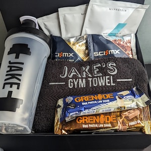 May include: A black gift box filled with gym-themed items, including a black shaker bottle with a white dumbbell and the name "JAKE" printed on it, a black towel with the text "JAKE'S GYM TOWEL", protein powder sachets, protein bars, and a grey weight plate.