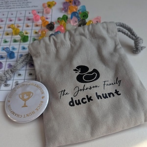 May include: A light grey drawstring bag with a black duck logo and the text "The Johnson Family duck hunt." Includes a white button with a gold trophy and the text "Duck Hunt Champion." Scattered are colourful plastic duck game pieces.
