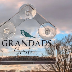 May include: A clear acrylic bird feeder with suction cups, designed to attach to a window. The feeder has a small bird silhouette and the text "Grandads Garden".