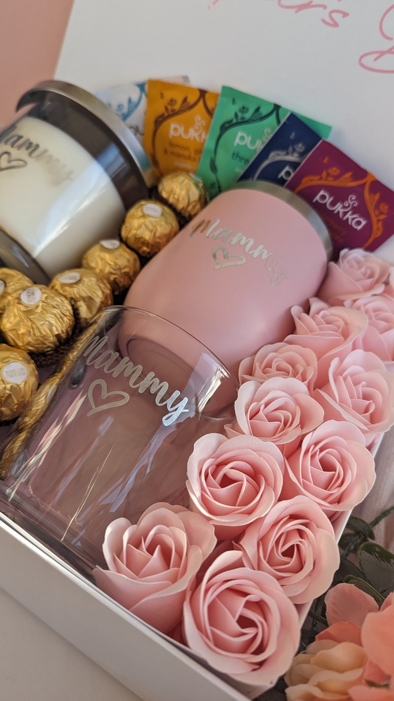 May include: A gift box filled with a pink tumbler and a glass with the word "Mommy" on them, surrounded by pink roses and chocolate sweets.