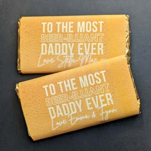 May include: Two yellow chocolate bar wrappers with a white background. The text on the wrappers reads "To the most beer-illiant daddy ever". One wrapper says "Love Stella-Mae" and the other says "Love Emma & Flynn".