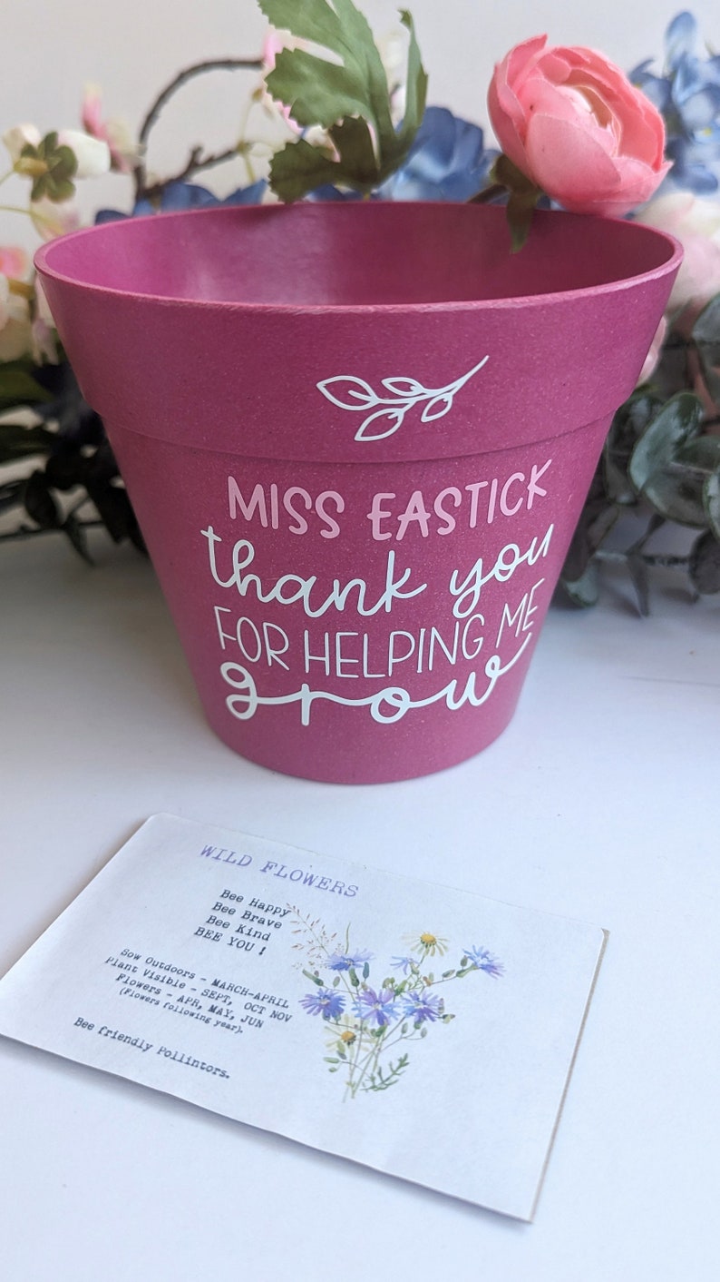 May include: A pink ceramic planter with the text "MISS EASTICK thank you FOR HELPING ME GROW" in white lettering. A small card with the text "WILD FLOWERS Bee Happy Bee Brave Bee Kind BEE YOU! Sow Outdoors - MARCH-APRIL Plant Visible - SEPT, OCT NOV Flowers - APR, MAY, JUN (Flowers following year). Bee friendly Pollintors." is in the foreground.