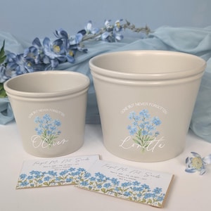 May include: Two beige ceramic flower pots with a floral design and the words "Gone But Never Forgotten." One pot has the name "Oliver," the other "Loretta." Two seed packets are in front.