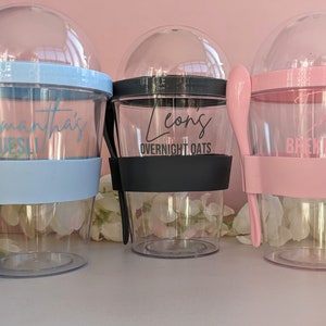 May include: Three clear plastic cups with lids and attached spoons. The cups are personalised with names and text. The cup on the left is light blue with the text "Samantha's Muesli". The middle cup is black with the text "Leon's Overnight Oats". The cup on the right is pink with the text "Emily's Breaky".