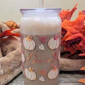 May include: A clear plastic tumbler filled with a creamy drink and a straw. The tumbler is decorated with gold and pink pumpkins, leaves, and the name "Daisy". The background includes autumn leaves and a soft blanket.