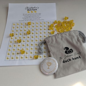 May include: A duck hunt game set featuring a printed game board, yellow duck-shaped game pieces, a drawstring bag with a duck logo, and a button that says "Duck Hunt Champion". The game board has the text "Christopher's duck hunt".