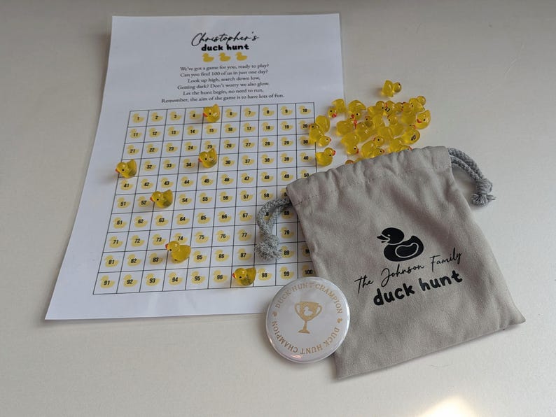 May include: A duck hunt game set featuring a printed game board, yellow duck-shaped game pieces, a drawstring bag with a duck logo, and a button that says "Duck Hunt Champion". The game board has the text "Christopher's duck hunt".