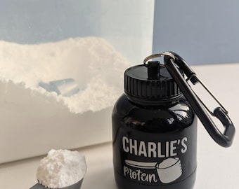 May include: Black plastic protein powder container with a carabiner clip. The container has a white label that says "CHARLIE'S protein" with a graphic of a scoop. A white scoop of powder is next to the container.