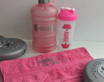 Puede incluir: Botella de agua rosa personalizada, botella de batido y toalla con el nombre Abbie y un gráfico de mancuernas. La toalla tiene el texto "Abbie's Gym Towel".