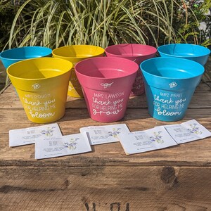 May include: Five colorful plant pots with personalized messages for teachers. The pots are yellow, blue, and pink. The messages say "Thank you for helping me bloom", "Thank you for helping me grow", and "Thank you for helping me bloom". The pots are sitting on a wooden crate with the words "Bleasdale Ltd Wholesale Chemists York" printed on the side.