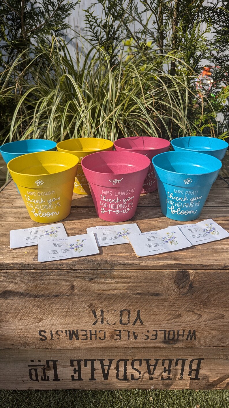 May include: Five colorful plant pots with personalized messages for teachers. The pots are yellow, blue, and pink. The messages say "Thank you for helping me bloom", "Thank you for helping me grow", and "Thank you for helping me bloom". The pots are sitting on a wooden crate with the words "Bleasdale Ltd Wholesale Chemists York" printed on the side.