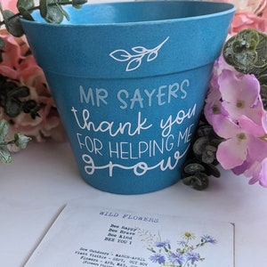 May include: A blue ceramic flower pot with white lettering that reads "Mr. Sayers thank you for helping me grow". The pot is surrounded by pink flowers.