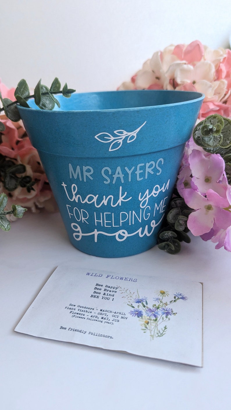 May include: A blue ceramic flower pot with white lettering that reads "Mr. Sayers thank you for helping me grow". The pot is surrounded by pink flowers.
