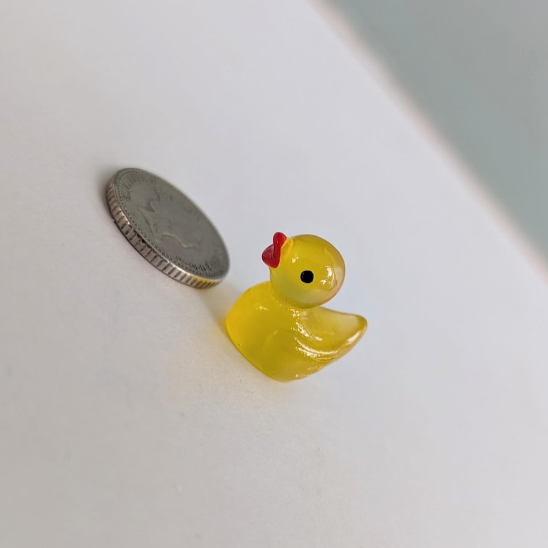 May include: A small, yellow rubber duck with a red bow, next to a silver coin. The duck has a glossy finish and a black eye. The coin is a quarter.