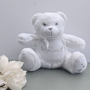 May include: White plush teddy bear with a white ribbon around its neck. The bear has a silver embroidered heart with wings and the name "Rose" on its chest. The bear's paws have three silver hearts on each.