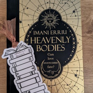 May include: A hardback book titled "Heavenly Bodies" by Imani Erriu, with a gold cover and a celestial design, and black details. A bookmark with a stack of books and the text "Heavenly Bodies" and "2026 Book Club" is placed on the book.