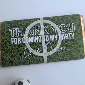 May include: A chocolate bar with a green grass-like background and the text "THANK YOU FOR COMING TO MY PARTY Love Owen" in white. Two footballs are on either side of the bar. The bar has gold foil edges.