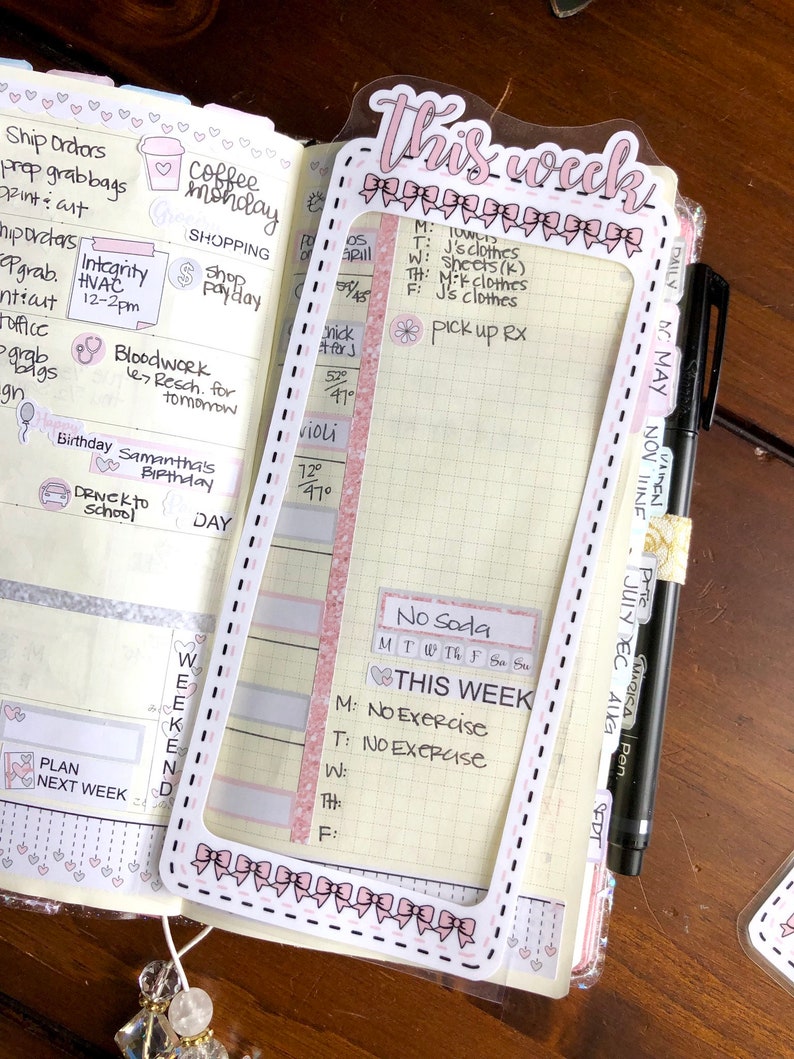 Op de afbeelding: Een roze en witte wekelijkse planner sticker met de tekst "This week" in roze cursief schrift. De sticker heeft een decoratieve rand van roze strikken en een wit frame. De sticker is geplaatst op een wekelijkse planner pagina met een rasterpatroon. De planner pagina heeft handgeschreven notities voor de week, waaronder "No Soda" en "No Exercise".