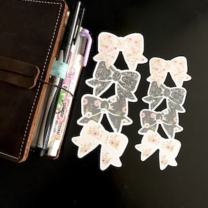May include: A set of six decorative bow stickers with floral and glitter designs. The stickers are white with pink and gray accents.