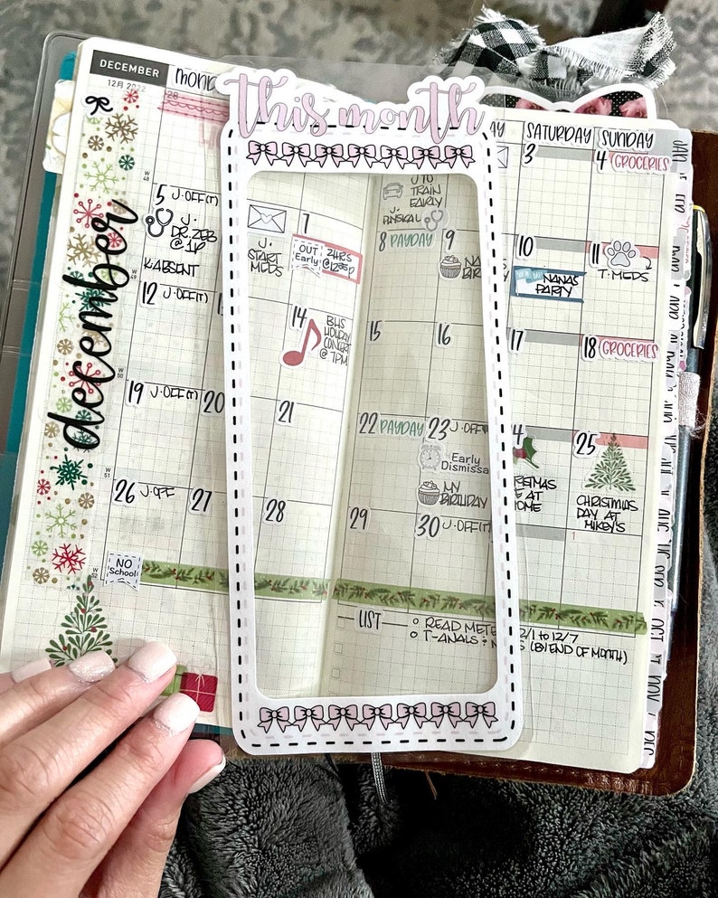 Op de afbeelding: Een transparante maandelijkse planner overlay van plastic met een witte rand en een roze titel "Deze maand". De overlay is geplaatst over een december-kalenderpagina met handgeschreven notities en stickers.