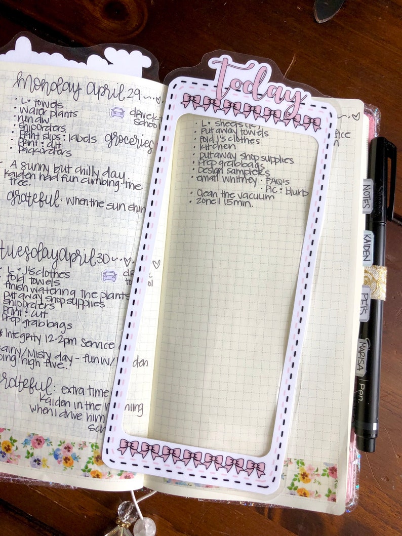 Op de afbeelding: Een close-up van een roze en witte planner met een doorzichtig plastic vel met het woord "Vandaag" erop. Het vel heeft een roze en wit strikdesign rond de randen. De planner heeft een rasterpatroon en is gevuld met handgeschreven notities.