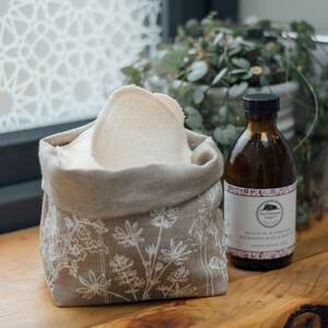 May include: A linen storage bag with a floral design, filled with reusable cotton rounds. A glass bottle of wild rose and geranium moisturizing bath and body soap sits next to the bag.