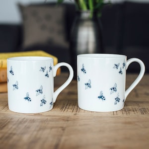 May include: Two white ceramic mugs with a blue bee pattern. The mugs have a white background and are decorated with blue bees in flight.