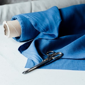 May include: A roll of blue linen fabric with a pair of silver scissors on top. The fabric is folded in half and the scissors are lying on top of the fabric.