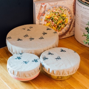 May include: Three reusable food covers made of natural fabric with a bee design. The covers are in different sizes and are shown on top of bowls.