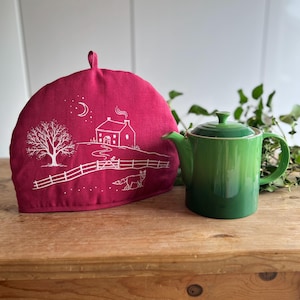 May include: A fuchsia tea cozy with a white landscape design featuring a house, tree, and fox. Beside it is a green ceramic teapot with a matching lid. The items are on a wooden surface.