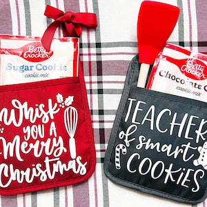 May include: Two red and black oven mitts with pockets. The red mitt has a white design that says "We Whisk You A Merry Christmas" and a whisk. The black mitt has a white design that says "Teacher of Smart Cookies" and a gingerbread man. Both mitts have a package of Betty Crocker cookie mix in the pocket.