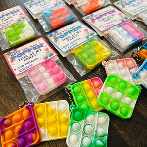 May include: A collection of colourful pop-it fidget toys with keychains. Each pop-it is packaged in a clear plastic bag with a printed tag that says "Hope you had a poppin' time at my party! Your friend, [name]"