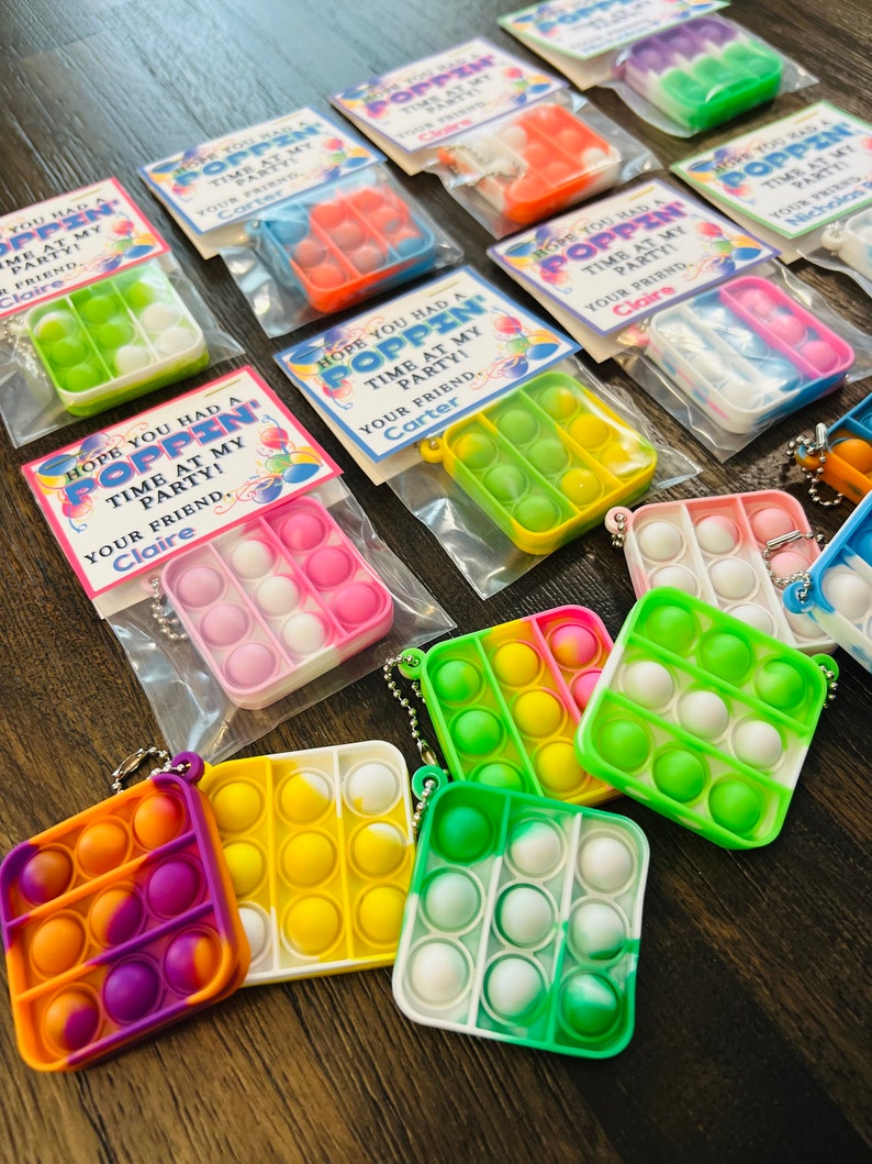 May include: A collection of colourful pop-it fidget toys with keychains. Each pop-it is packaged in a clear plastic bag with a printed tag that says "Hope you had a poppin' time at my party! Your friend, [name]"