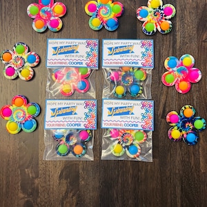 Pode incluir: Spinners fidget em forma de flor em várias cores, incluindo padrões tie-dye, são exibidos em uma superfície de madeira. Alguns spinners estão embalados em sacos transparentes com um cartão que diz "HOPE MY PARTY WAS Spinning WITH FUN! YOUR FRIEND, COOPER."
