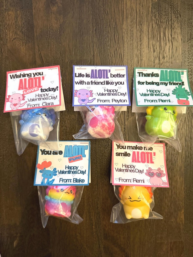 May include: Six Valentine's Day gifts featuring axolotl-themed toys in clear plastic bags. Each bag has a colorful card with a Valentine's Day message and the word "ALOTL". The axolotls are various colors, including blue, pink, green, rainbow, and yellow.