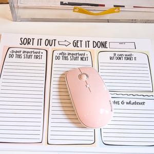 May include: A white desk pad with a colorful design and the text "SORT IT OUT GET IT DONE". The pad is divided into three sections with lined spaces for writing. A pink wireless mouse is on the pad.