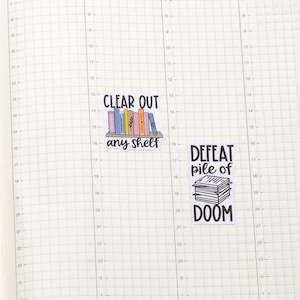 May include: Two colourful stickers with book-themed sayings. The first sticker says "Clear out any shelf" and features a bookshelf with colourful books. The second sticker says "Defeat pile of doom" and features a stack of books.