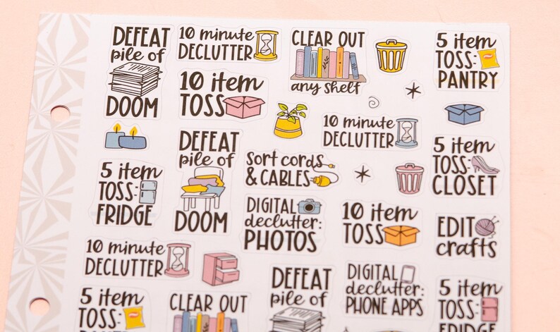 May include: A sheet of colourful stickers with various decluttering tasks. The stickers feature illustrations of a bin, a pile of clothes, a box, a cord, a camera, and a phone. The text on the stickers includes "DEFEAT pile of DOOM", "10 minute DECLUTTER", "10 item TOSS", "5 item TOSS", "CLEAR OUT any shelf", "Sort cords & CABLES", "DIGITAL declutter: PHOTOS", "DIGITAL declutter: PHONE APPS", "EDIT crafts", "5 item TOSS: PANTRY", "5 item TOSS: CLOSET", "5 item TOSS: FRIDGE", "5 item TOSS: FRIDGE".