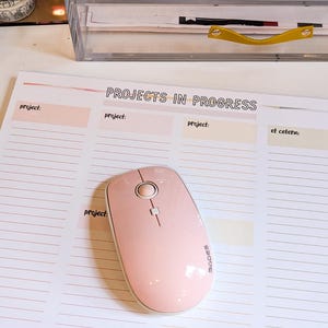 May include: A pink wireless mouse with a white scroll wheel and a white button on top. The mouse is on a white desk mat with the words "Projects in Progress" written in black at the top. The desk mat has lines for writing down projects.