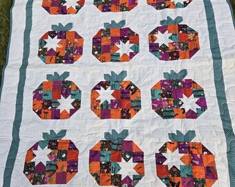 May include: A handmade quilt featuring a repeating pattern of colorful pumpkin shapes with star-shaped cutouts. The pumpkins are made from patchwork squares in orange, purple, and teal. The quilt has a white background with a teal border.