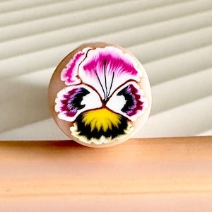 May include: A close-up of a small, round, hand-painted polymer clay pendant featuring a pansy flower with pink, purple, yellow, and black details.
