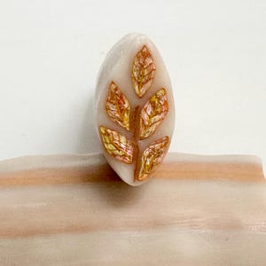 May include: An oval-shaped, off-white bead with a leaf design. The leaf design features a brown stem and five leaves with gold and pink iridescent accents. The bead is set on a light brown surface.