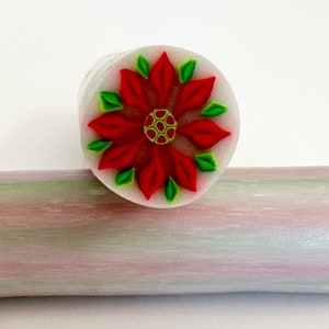May include: A polymer clay cane with a poinsettia design. The flower has red petals and green leaves, with a green and yellow center. The cane is white with a red and green rolling pin in the background.
